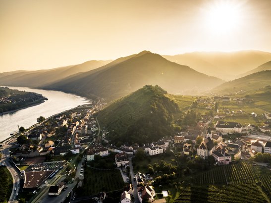Ein atemberaubender Blick auf ein Dorf, umgeben von Weinbergen und Bergen, mit einem glitzernden Fluss im Vordergrund.