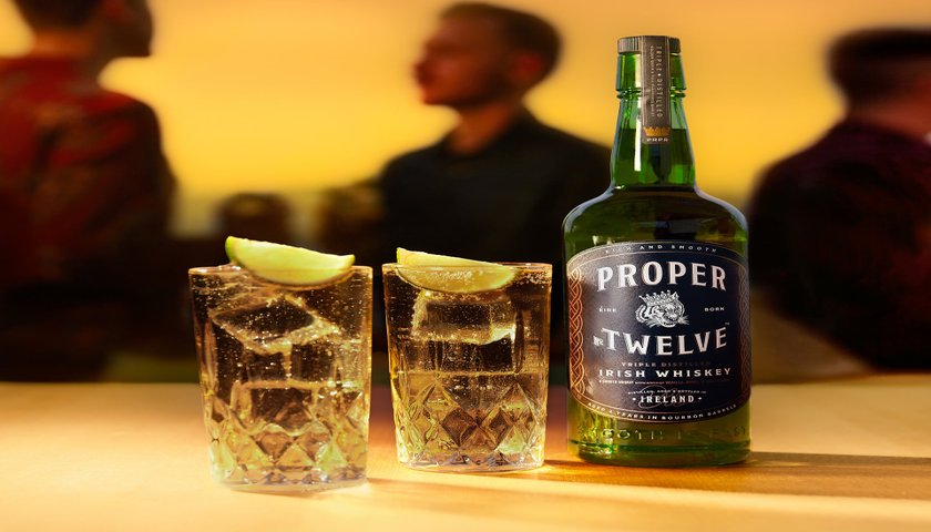 Zwei Gläser mit Eis und Limettenscheiben stehen neben einer Flasche Proper Twelve Irish Whiskey auf einem Tisch.
