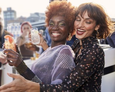 Zwei Frauen lachen fröhlich, während sie Cocktails in der Hand halten, umgeben von einer festlichen Atmosphäre.