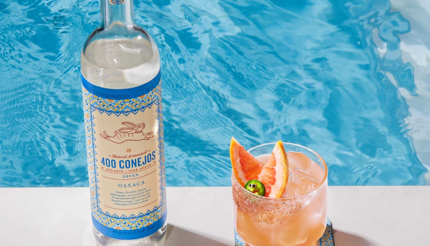 Eine Flasche Mezcal 400 Conejos steht neben einem Cocktailglas mit einer fruchtigen Garnitur am Rand eines blauen Pools.