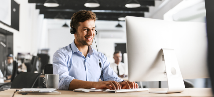 Ein junger Mann mit Headset lächelt, während er an einem Computer in einem modernen Büro arbeitet.