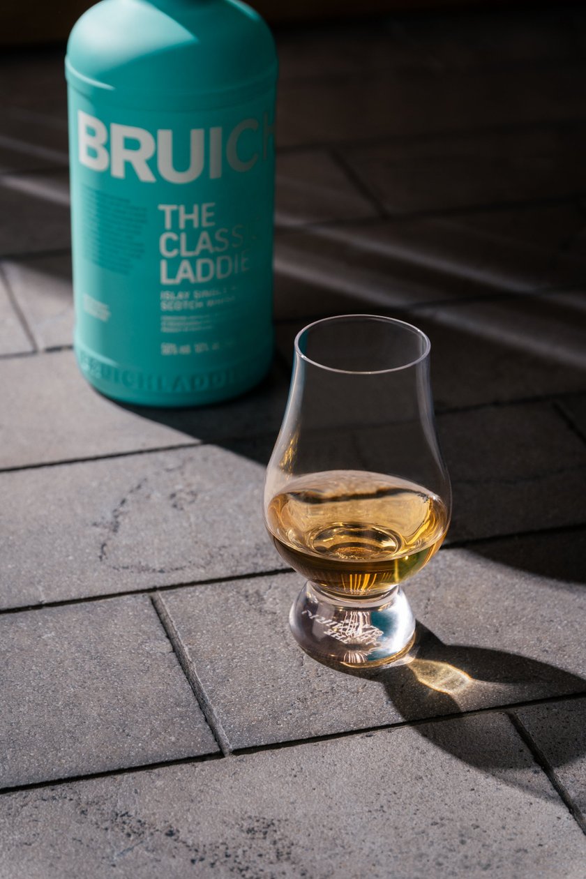 Ein elegantes Glas mit goldenem Whisky steht auf einem grauen Steinboden, während eine Flasche Bruichladdich im Hintergrund sichtbar ist.