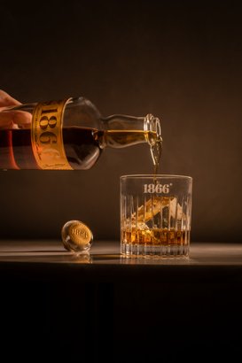 Ein elegantes Glas wird mit einem bernsteinfarbenen Getränk gefüllt, während die Flasche 1866 im sanften Licht glänzt.