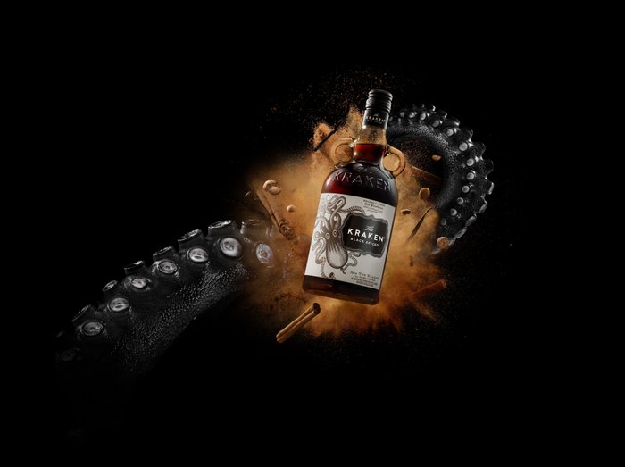 Eine Flasche Kraken Rum schwebt vor einem schwarzen Hintergrund, umgeben von einem Tentakel und einer Explosion von Gewürzen.
