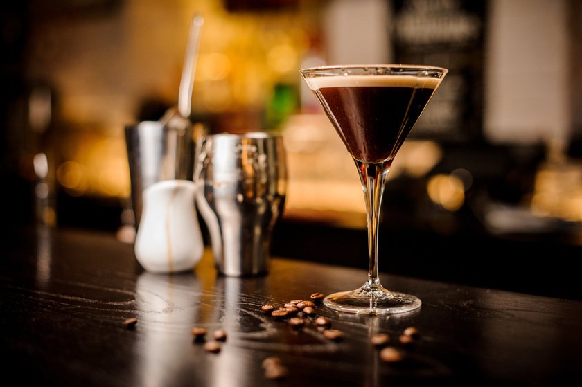 Ein eleganter Cocktail in einem Martiniglas steht auf einem dunklen Tisch, umgeben von Kaffeebohnen und Barutensilien.