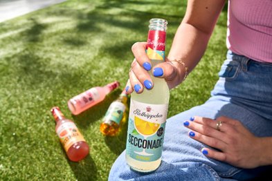 Eine Person mit blauen Nägeln hält eine Flasche Zitronensecconade in der Hand, während sie auf grünem Gras sitzt.