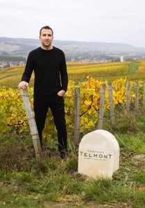 In einem malerischen Weinberg steht ein Mann in schwarzer Kleidung neben einem markanten Stein mit der Aufschrift  'Champagne Telmont'.