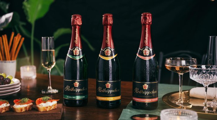 Drei Flaschen Sekt stehen auf einem Tisch, umgeben von Gläsern und Snacks, die eine festliche Atmosphäre schaffen.