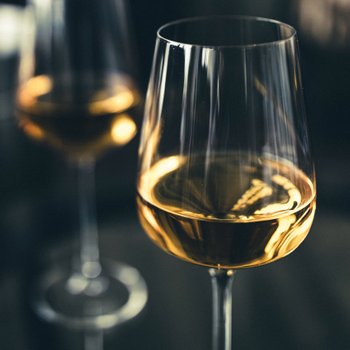 Ein elegantes Weinglas mit goldfarbenem Inhalt steht im Vordergrund, während ein verschwommenes Glas im Hintergrund sichtbar ist.
