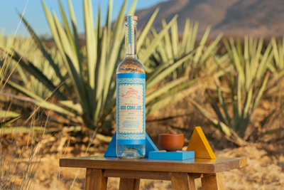 In einer ländlichen Umgebung steht eine Flasche Mezcal auf einem Tisch, umgeben von Agaven und einer warmen, einladenden Atmosphäre.