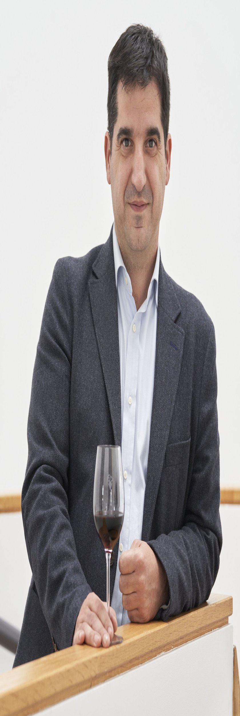 Ein Mann in einem grauen Blazer steht an einem Holzgeländer und hält ein Glas Rotwein in der Hand.