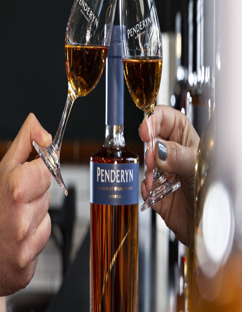 Zwei Gläser mit Penderyn Whisky stoßen an, während eine Flasche im Hintergrund steht, die die goldene Farbe des Getränks reflektiert.