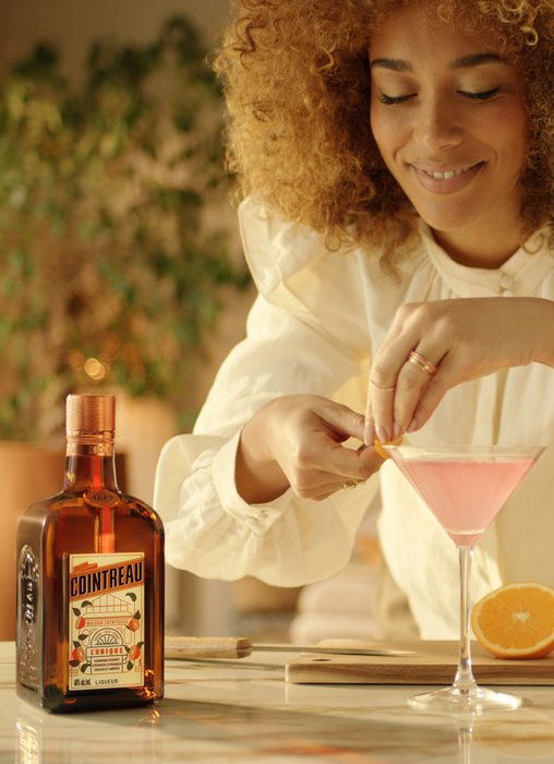 Eine fröhliche Frau mit lockigem Haar dekoriert einen rosa Cocktail mit einer Orangenscheibe in einer stilvollen Bar.