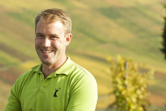 Ein lächelnder Mann in einem leuchtend grünen Poloshirt steht vor einer malerischen, hügeligen Landschaft mit sanften Farben.