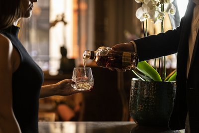 In einem luxuriösen Setting wird einer Frau von einem Mann ein Getränk in ein Glas eingeschenkt, umgeben von Pflanzen.
