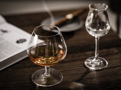 Ein elegantes Glas mit goldfarbenem Getränk steht auf einem Holztisch, daneben ein weiteres Glas mit klarem Inhalt.