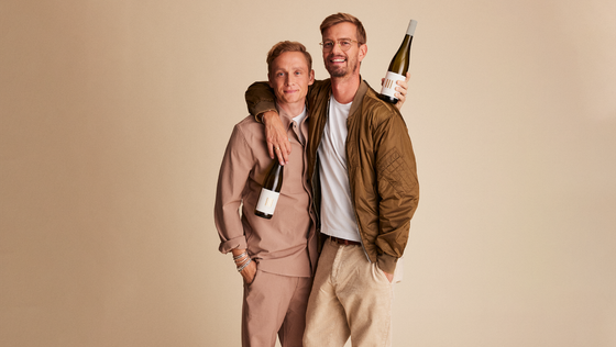 Zwei Männer stehen eng umarmt vor einem neutralen Hintergrund und halten Flaschen Wein in der Hand, lächelnd und glücklich.