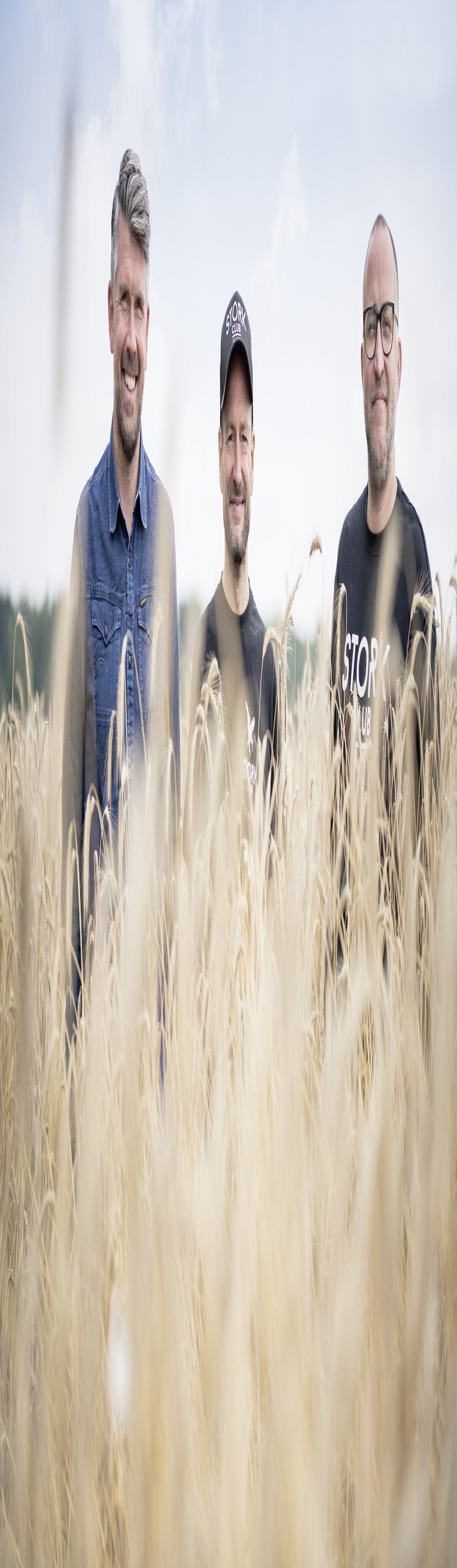Drei Männer stehen lächelnd in einem goldenen Getreidefeld, umgeben von hohen Halmen unter einem bewölkten Himmel.