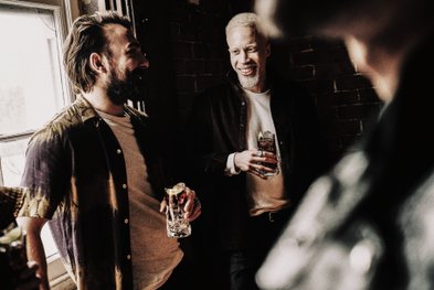 Zwei Männer stehen in einem gemütlichen Raum, lachen und halten Gläser mit Getränken in der Hand.