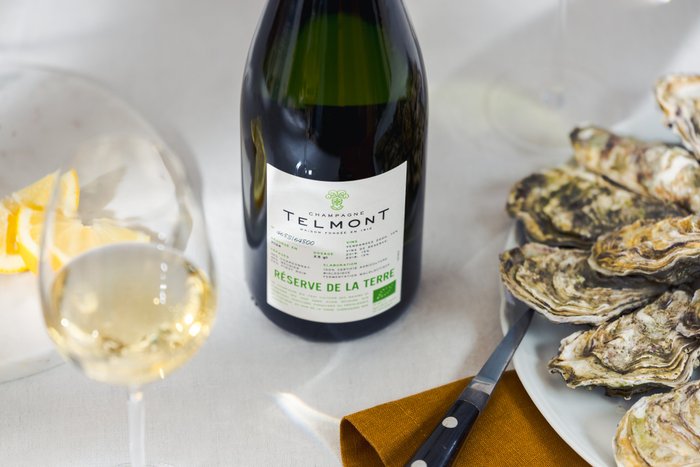 Ein elegantes Arrangement mit einer Flasche Telmont Champagner, einem Glas und einem Teller voller Austern, dekoriert mit Zitrone.