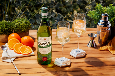 Eine Flasche Carpano Bianco steht auf einem Holztisch, umgeben von frischen Orangen und zwei Gläsern mit Eis.