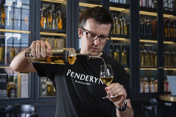 Ein Mann mit Brille gießt sorgfältig einen goldenen Likör in ein Glas mit dem Schriftzug Penderyn.